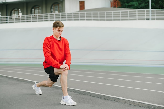 Coureur dans un sweat-shirt rouge se réchauffe avant de courir sur la piste et sur le terrain de sport regarde vers le bas Un sportif sérieux réchauffe ses muscles avant une séance d'entraînement