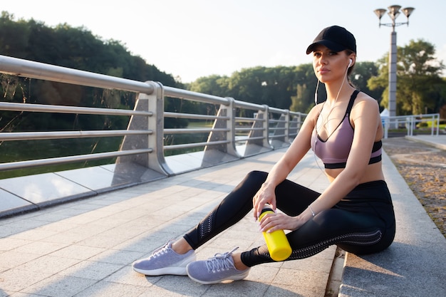 Coureur de jeune femme se reposant après une séance d'entraînement le matin ensoleillé. Modèle de fitness féminin assis dans la rue le long de l'étang de la ville. Jogger féminin prenant une pause de l'entraînement en cours d'exécution