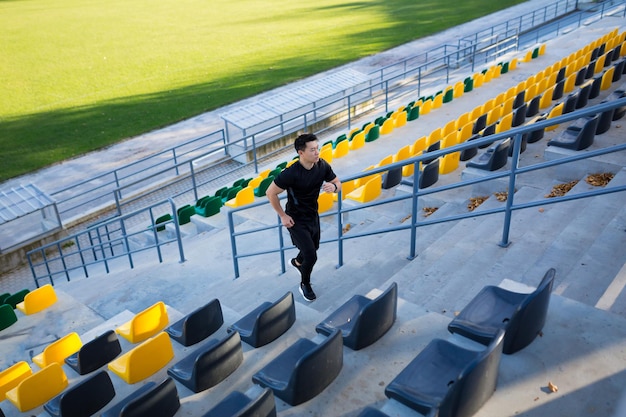 Coureur masculin asiatique courant autour du stade urbain moderne dans les escaliers Cardio jogging matinal escalade homme escalier jogging en plein air. entraînement sportif homme athlète par étapes, en ville. Mode de vie sain de remise en forme active