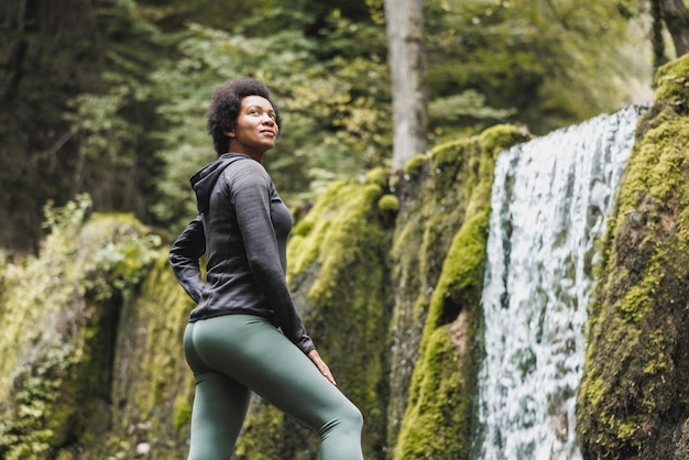 Une coureuse afro-américaine mature se détend après avoir fait du jogging près de la cascade des montagnes dans la nature.