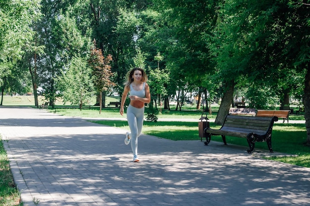 Courir une jeune femme sportive aux cheveux longs et lâche qui s'échauffe en s'étirant dans des leggings et des baskets gris avec une montre dans une journée d'été ensoleillée près d'un banc et d'une poubelle parmi les arbres verts du parc