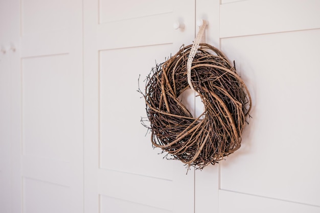 Couronne de saule suspendue marron tissée en osier avec dentelle décorative en ruban blanc Armoire blanche en arrière-plan Décoration traditionnelle de vacances ethniques saisonnières Symbole de salutation de conception sur la poignée de porte