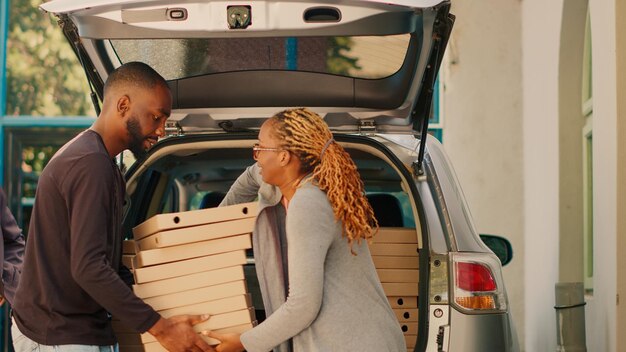 Courrier afro-américain donnant des tas de boîtes à pizza à diverses personnes debout près de l'entrée du bâtiment. Employé de livraison de nourriture livrant des piles de colis de repas dans le coffre de la voiture. Prise de vue au trépied.