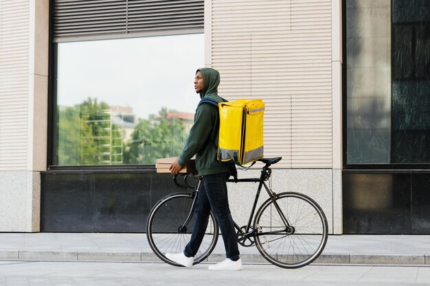 Un courrier afro-américain à vélo Un homme tient des boîtes de pizza devant des bâtiments modernes en arrière-plan