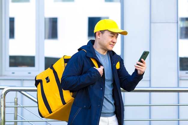 Courrier alimentaire masculin asiatique utilisant un mobile à l'aide d'une application de téléphone portable sur un smartphone tout en se tenant dans la rue de la ville avec un sac à dos thermique isolé livrant de la nourriture. à la recherche de l'adresse du client. acheter une carte en ligne