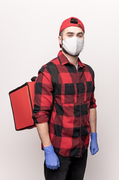 Courrier contemporain en vêtements de travail et masque de protection et gants portant un grand sac rouge avec des commandes de clients de café sur son dos