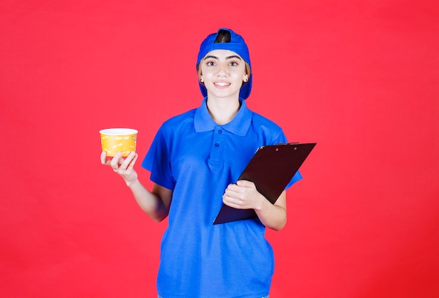 Courrier féminin en uniforme bleu tenant une tasse de nouilles jaunes et un dossier noir.