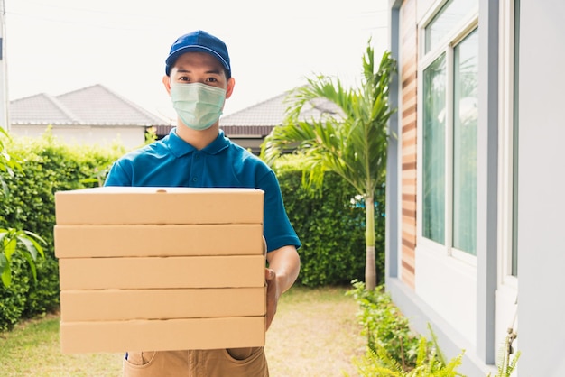 Courrier de jeune livreur asiatique envoyant et tenant une pizza de restauration rapide en boîte en uniforme, il protège le client du service de masque facial à la porte de la maison, sous couvre-feu coronavirus pandémique de quarantaine COVID-19