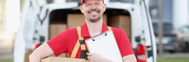 Le courrier masculin en uniforme tient le presse-papiers et la boîte en carton en arrière-plan du camion avec des boîtes