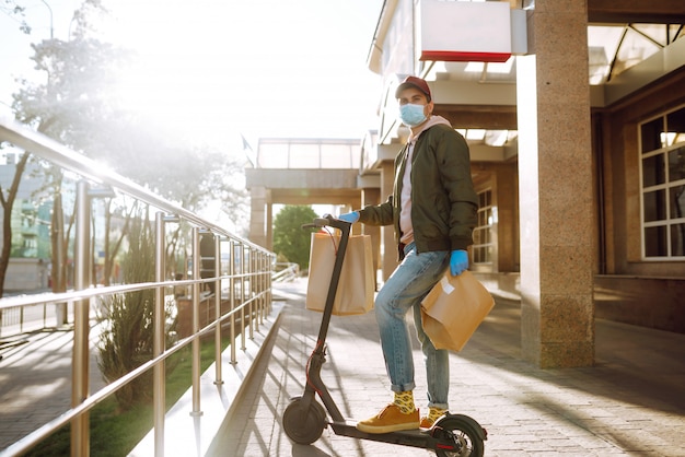 Courrier en masque de protection et gants médicaux sur un scooter livre un paquet de papier kraft avec de la nourriture Service de livraison en quarantaine, éclosion de maladie, pandémie de coronavirus covid-19.