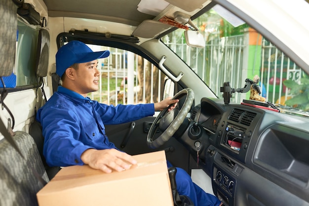 Courrier positif souriant en uniforme de conduite de camion de livraison avec boîte en carton sur le siège avant