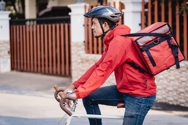 Courrier en uniforme rouge avec une boîte de livraison à l'arrière faisant du vélo et regardant sur le téléphone portable pour vérifier l'adresse pour livrer la nourriture au client. Courrier à vélo livrant de la nourriture dans la ville.