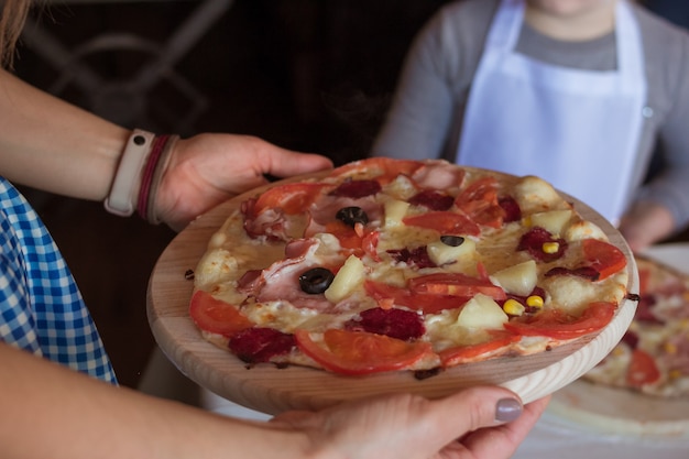 Cours de cuisine, culinaire. Mains de femme tenant une pizza cuite au four avec des tomates, du fromage, du bacon, du salami, du maïs et des olives sur une planche à découper en bois ronde. Enfants préparant la nourriture, repas dans la cuisine