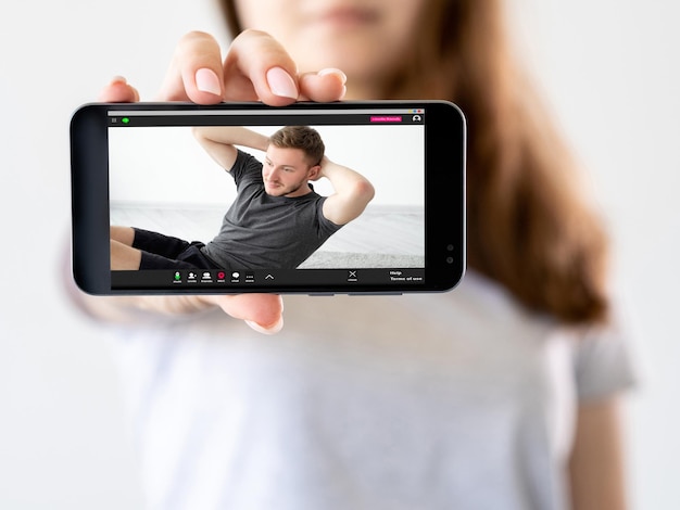 Cours de fitness virtuel Formation en ligne Tutoriel d'entraînement Main de fille montrant un programme vidéo de sport sur téléphone avec un homme athlétique faisant des redressements assis sur écran isolé sur neutre défocalisé