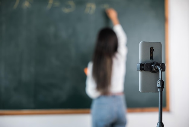 Cours en ligne de retour à l'école. Une enseignante asiatique enseigne à bord tout en ayant une conférence au tableau noir en ligne via un smartphone dans la salle de classe, une femme debout devant la classe en direct un appel vidéo en ligne sur un téléphone mobile intelligent