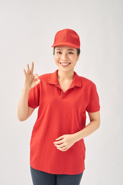 Coursier de jeune fille heureuse, portant l'uniforme de casquette regardant la caméra montrant le signe ok recommande un service de livraison rapide sur fond blanc.