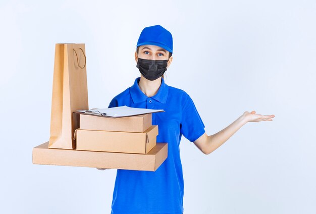 Coursière en masque et uniforme bleu tenant un sac à provisions en carton et plusieurs boîtes et pointant vers quelqu'un.