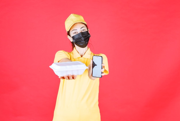 Photo coursière en uniforme jaune et masque noir tenant un colis à emporter et prenant les commandes via smartphone