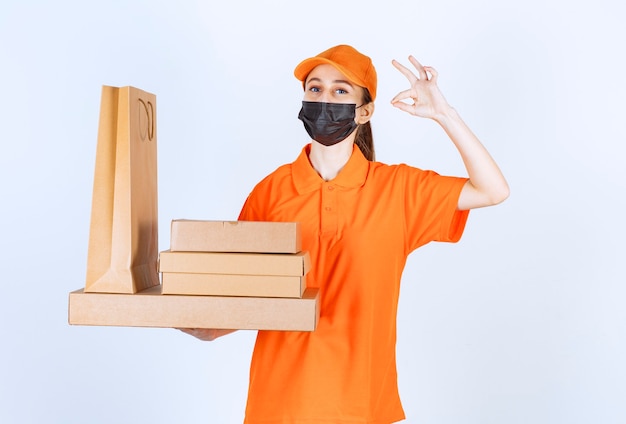 Coursière en uniforme jaune et masque noir tenant plusieurs colis en carton et un sac à provisions et montrant un signe positif de la main.