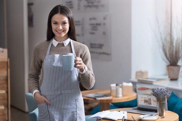 Court repos. Charmante belle femme souriante et debout dans un café tout en tenant une tasse de thé.