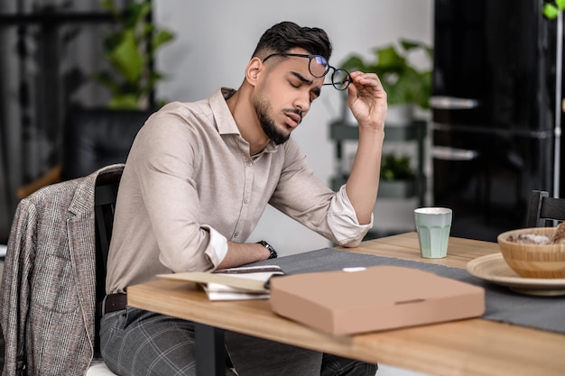 Courte pause. Séduisant jeune homme barbu aux yeux fermés enlevant des lunettes assis à table à la maison