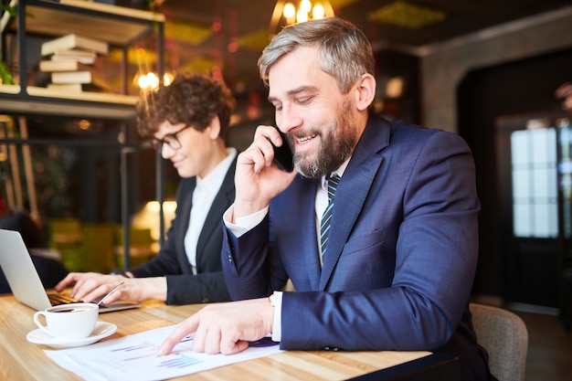 Courtier prospère en costume élégant analysant des graphiques sur des papiers tout en parlant à un collègue ou à un client sur smartphone