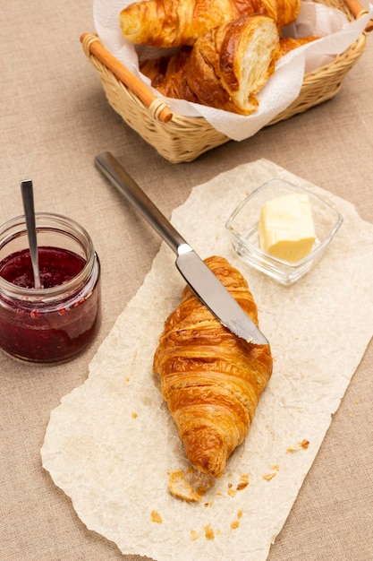 Couteau de cuisine et bol avec beurre, croissants sur papier. Confiture en bocal. Linge de jute sur table. Vue de dessus.