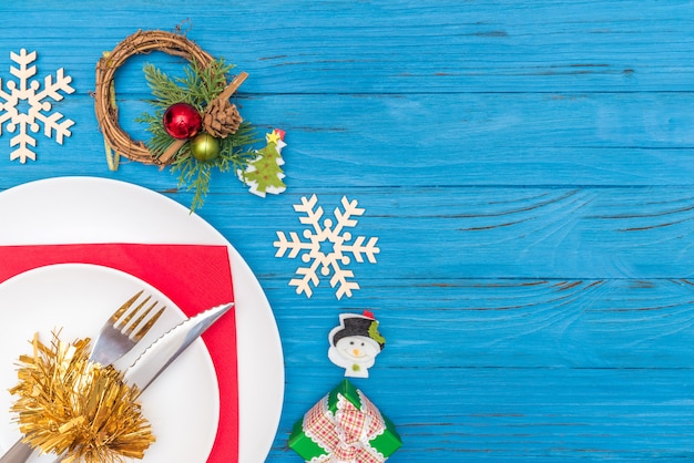 Un couteau et une fourchette en plaque blanche sur une serviette rouge décorée d'une couronne de Noël en flocons de neige en bois et d'une boîte-cadeau