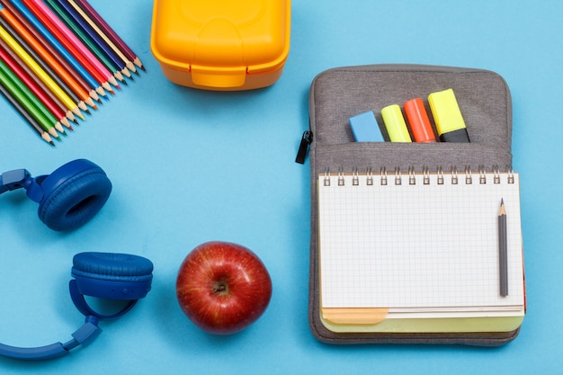 Écouteurs, crayons de couleur, boîte à lunch, pomme, cahier ouvert sur étui à crayons avec feutres de couleur et marqueur sur fond bleu. Vue de dessus. Retour au concept de l'école. Fournitures scolaires.