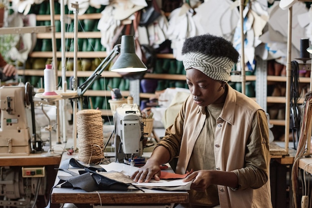 Une couturière africaine travaillant avec des motifs et des morceaux de tissus pour coudre de nouveaux vêtements à table dans l'usine