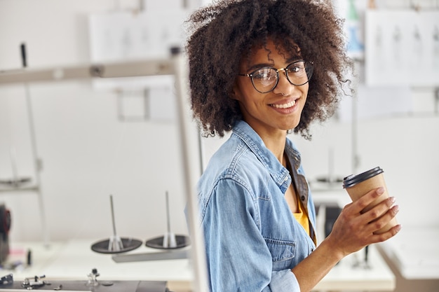 La couturière afro-américaine avec des lunettes et une boucle d'oreille dans le nez tient une tasse près de la machine à coudre dedans