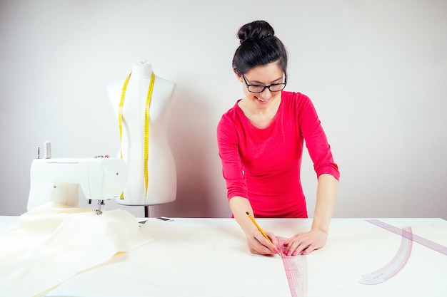 Couturière de belle fille avec des lunettes travaillant avec un crayon et des motifs Fond de mannequin avec machine à coudre à ruban jaune et fond blanc