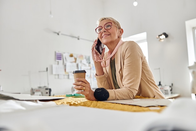 Une couturière féminine avec une tasse en papier et un oreiller à épingles parle sur un téléphone portable s'appuyant sur une table à découper