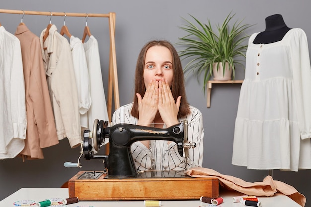 Une couturière surprise et choquée posant sur son lieu de travail cousant des vêtements a fait une erreur de couturière en faisant un mauvais point de matériau gâté pour une robe couvrant la bouche avec les mains