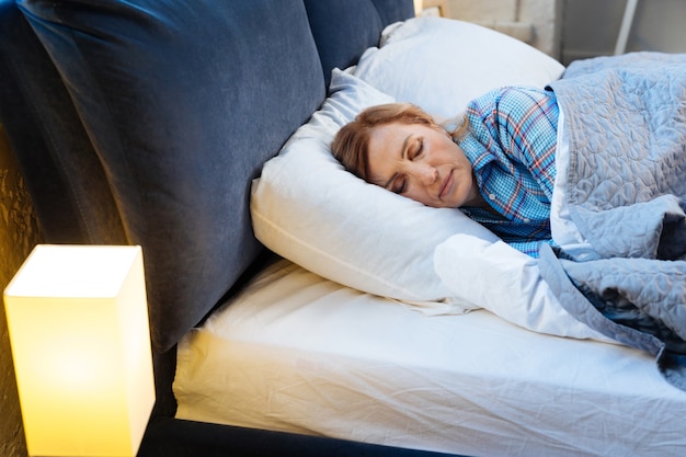 Couvert et chaud. Femme aux cheveux clairs adulte allongée les yeux fermés recouverte d'une couverture chaude tout en portant un pyjama lumineux