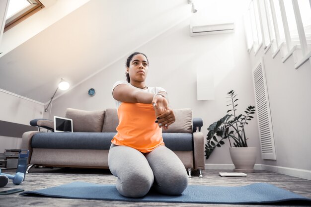 Couvert de tatouages. Femme afro-américaine aux cheveux attachés faisant un exercice spécial pour les paumes alors qu'il était assis sur le tapis de yoga