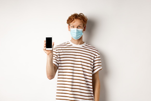 Covid-19, concept de médecin en ligne. Homme rousse souriant dans un masque facial montrant l'écran du smartphone, debout sur fond blanc.