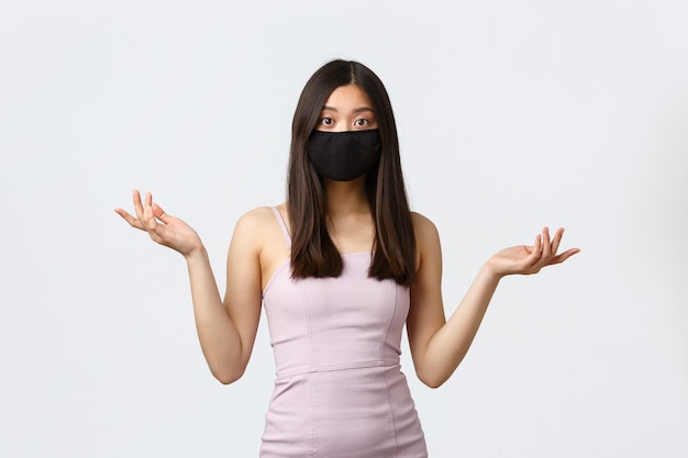 Covid-19, concept de quarantaine et de loisirs à distance sociale. Femme asiatique confuse en robe de soirée et masque facial, ne peut pas comprendre ce qui s'est passé