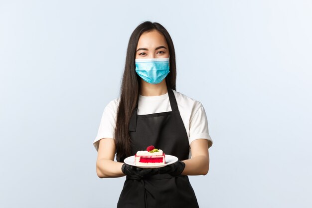 Covid-19, distanciation sociale, petite entreprise de café et concept de prévention des virus. Belle serveuse asiatique souriante, barista en masque médical et gants, servant un délicieux gâteau au client sur une assiette.