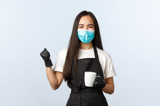 Covid-19, distanciation sociale, petite entreprise de café et concept de prévention des virus. La fille a finalement fait un délicieux café alors qu'elle commençait à travailler dans un café, une pompe à poing, se réjouissant, tenant une tasse avec une boisson.