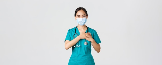 Covid-19, maladie à coronavirus, concept des travailleurs de la santé. Femme médecin asiatique bienveillante et attentionnée, médecin portant un masque médical et des gants, tenant les mains sur le cœur et souriant, fond blanc