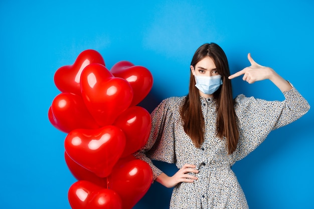 Covid-19, quarantaine et concept de santé. Jeune femme impertinente en masque médical, pointant du doigt les ballons coeurs rouges et fronçant les sourcils, déçue de la Saint-Valentin, fond bleu