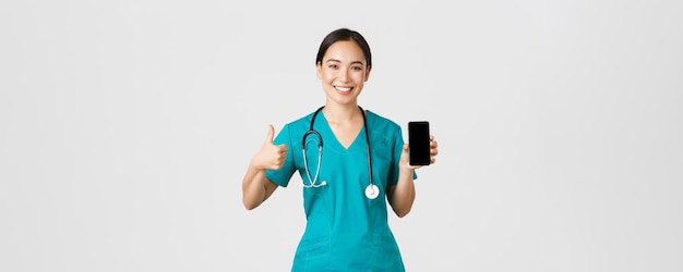 Covid-19, travailleurs de la santé et concept de médecine en ligne. Portrait d'une jeune femme médecin asiatique, médecin en gommage montrant le pouce levé et l'écran du smartphone, promouvoir l'application, fond blanc.