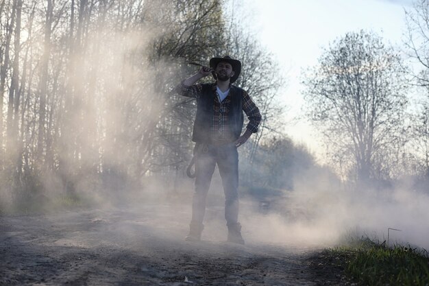 Un cowboy dans un chapeau et un gilet se tient dans une épaisse fumée sur la route