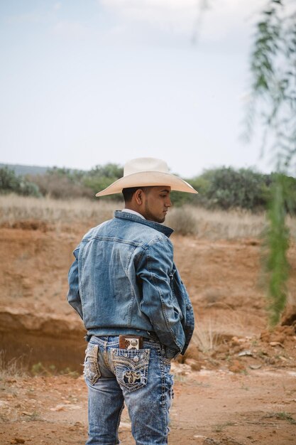 Cowboy portant des jeans et un chapeau debout sous le ciel avec des nuages moelleux travaillant dans une ferme