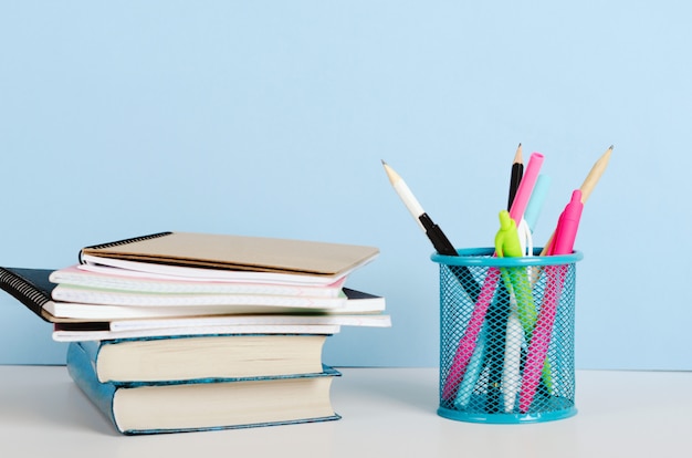 Crayons multicolores, stylos dans un support et livres avec des cahiers sur un tableau blanc sur fond bleu, lieu de travail de bureau. Copiez l'espace.