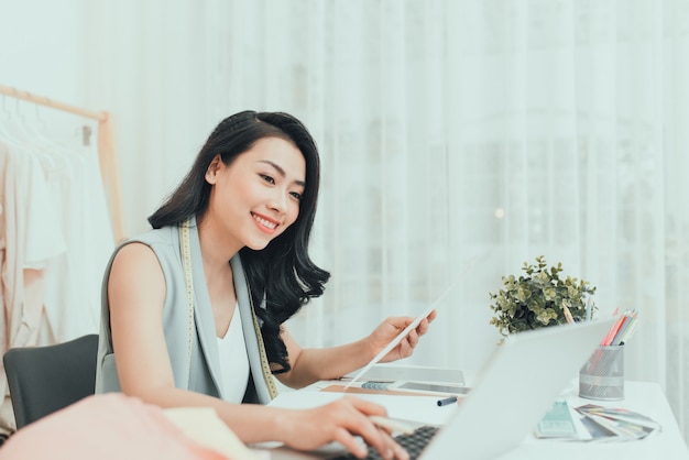 Créateur de mode au travail. Heureuse jeune femme asiatique dessinant assise sur son lieu de travail dans un atelier de mode