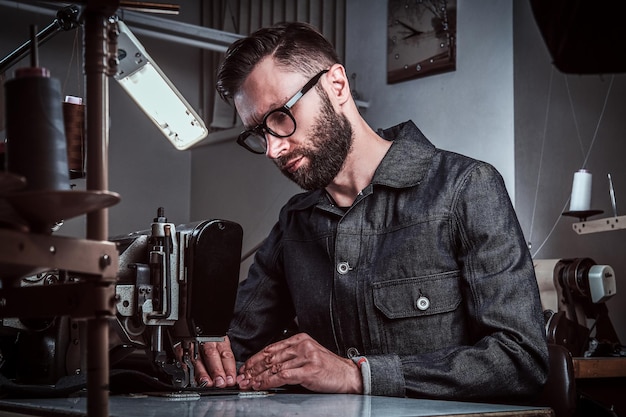 Le créateur de mode coud des vêtements à la machine à coudre dans son atelier. Il porte des lunettes et un jean. Il y a beaucoup de bobines de fils à l'arrière-plan.