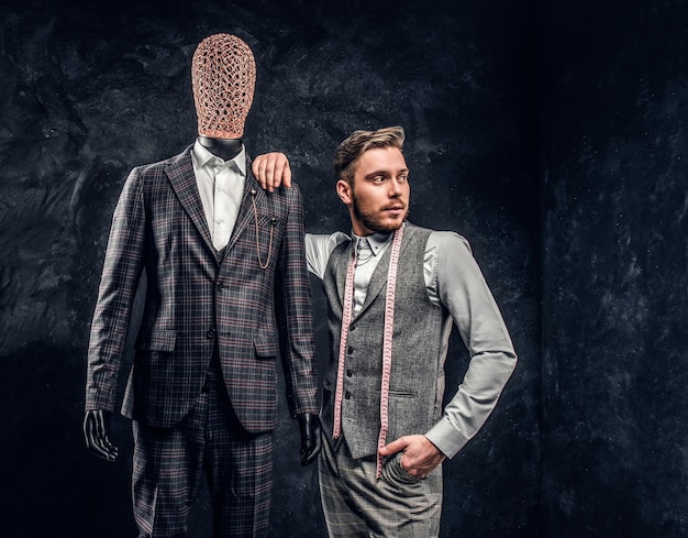 Un créateur de mode élégamment habillé posant à côté d'un mannequin en costume exclusif pour hommes sur mesure dans un studio de tailleur sombre