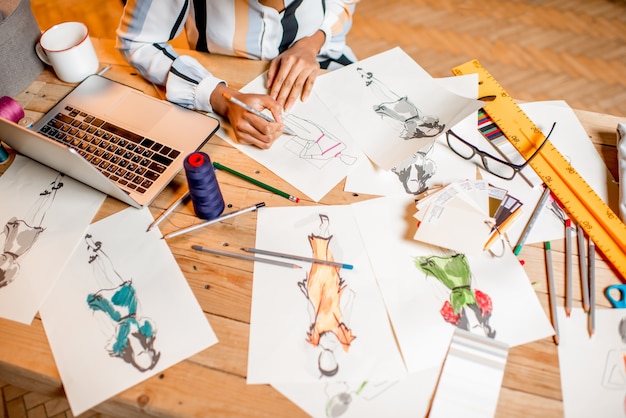 Créateur de mode esquissant des dessins de vêtements à table avec des outils de couture et un ordinateur portable